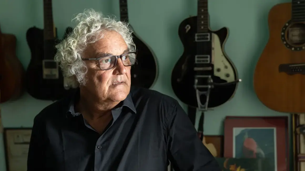 Massimo Bubola, con i suoi capelli ricci e bianchi, guarda pensieroso tra le chitarre appese al muro, testimoni silenziose delle sue canzoni.