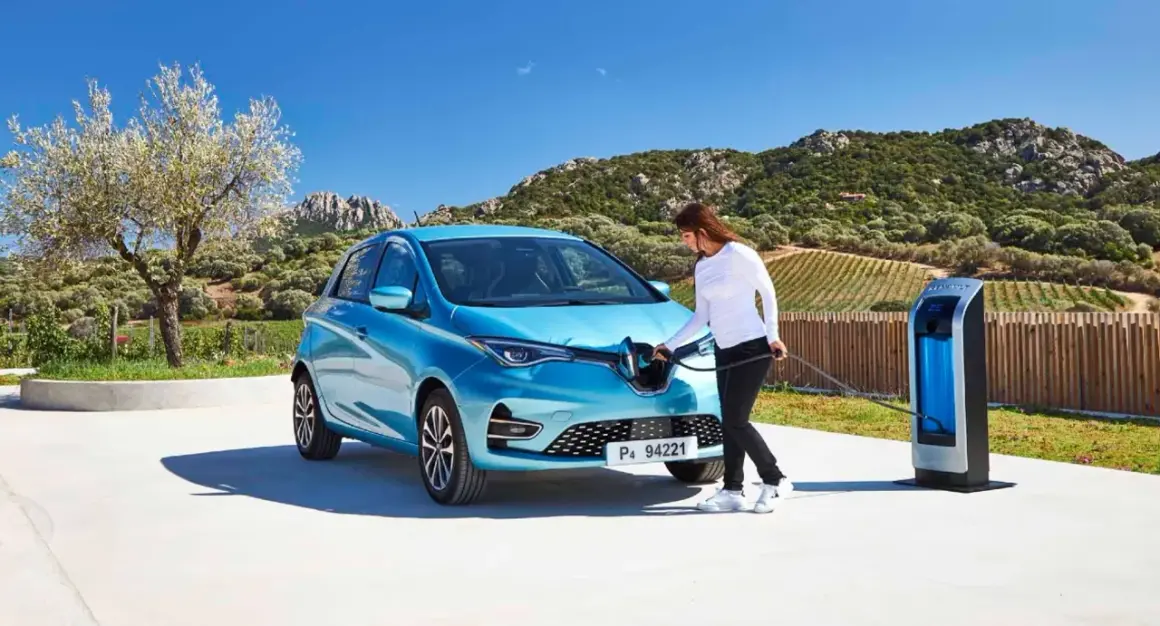 Une femme branche sa Renault Zoé bleue à une borne de recharge. Le temps de recharge zoe 22kw est rapide, parfait pour profiter du paysage.