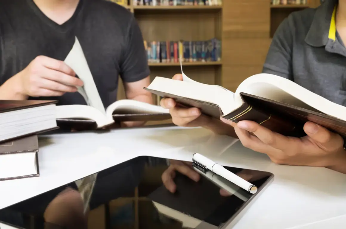 Estudiantes consultan libros y tablet en biblioteca, preparándose para las competencias específicas LOMLOE Andalucía.