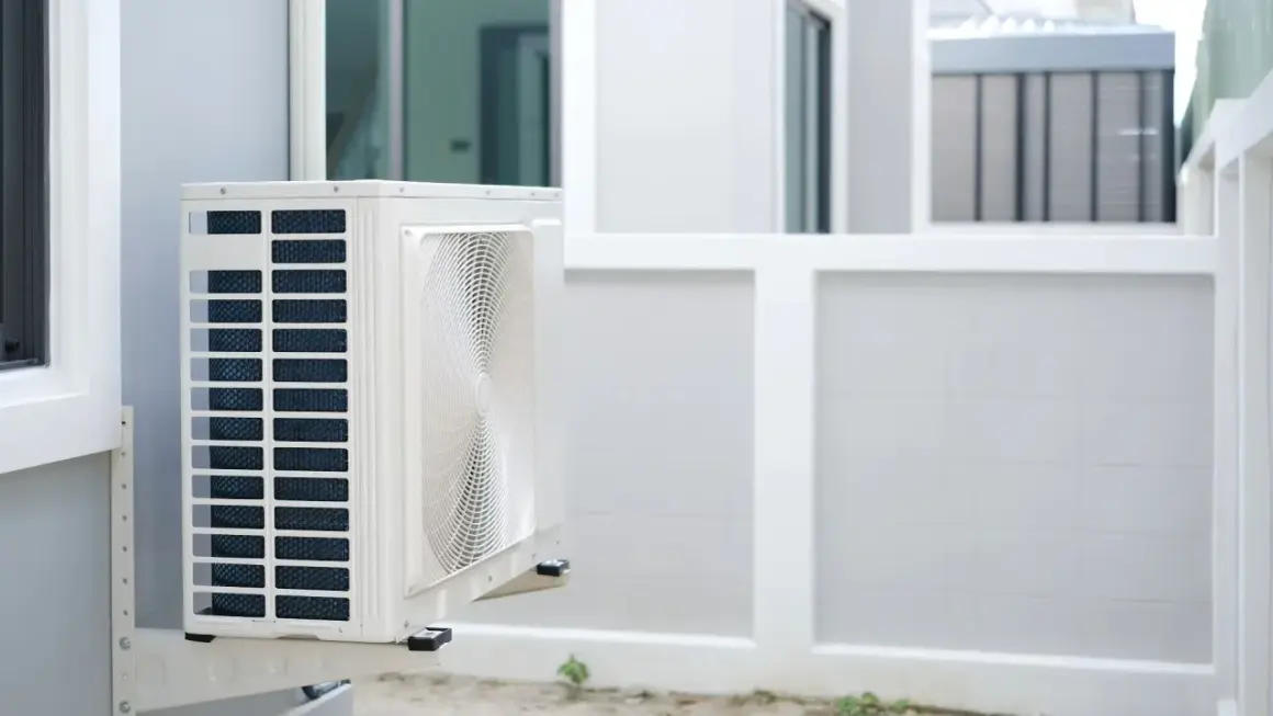 Unité extérieure d'une pompe à chaleur appartement, fixée sur un mur blanc. L'appareil est blanc avec des grilles bleues et un ventilateur visible.