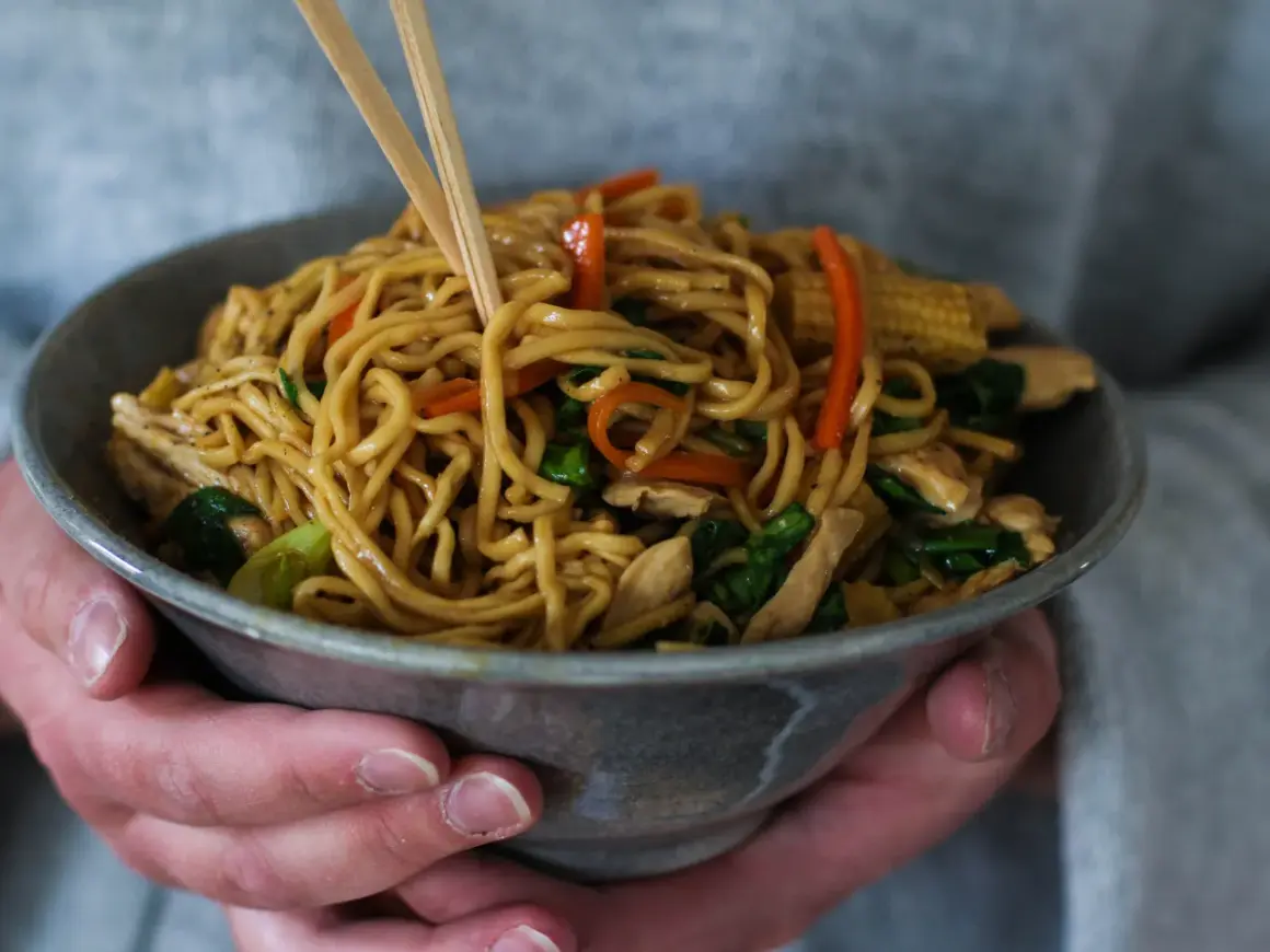 En skål med lo mein-nudlar, kyckling och grönsaker, lyfts med ätpinnar.