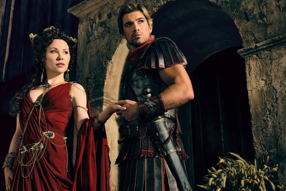 Die Besetzung von Spartacus: Ashur und Lucretia, in prachtvollen Gewändern und Rüstungen, halten Händchen vor einer antiken Kulisse.