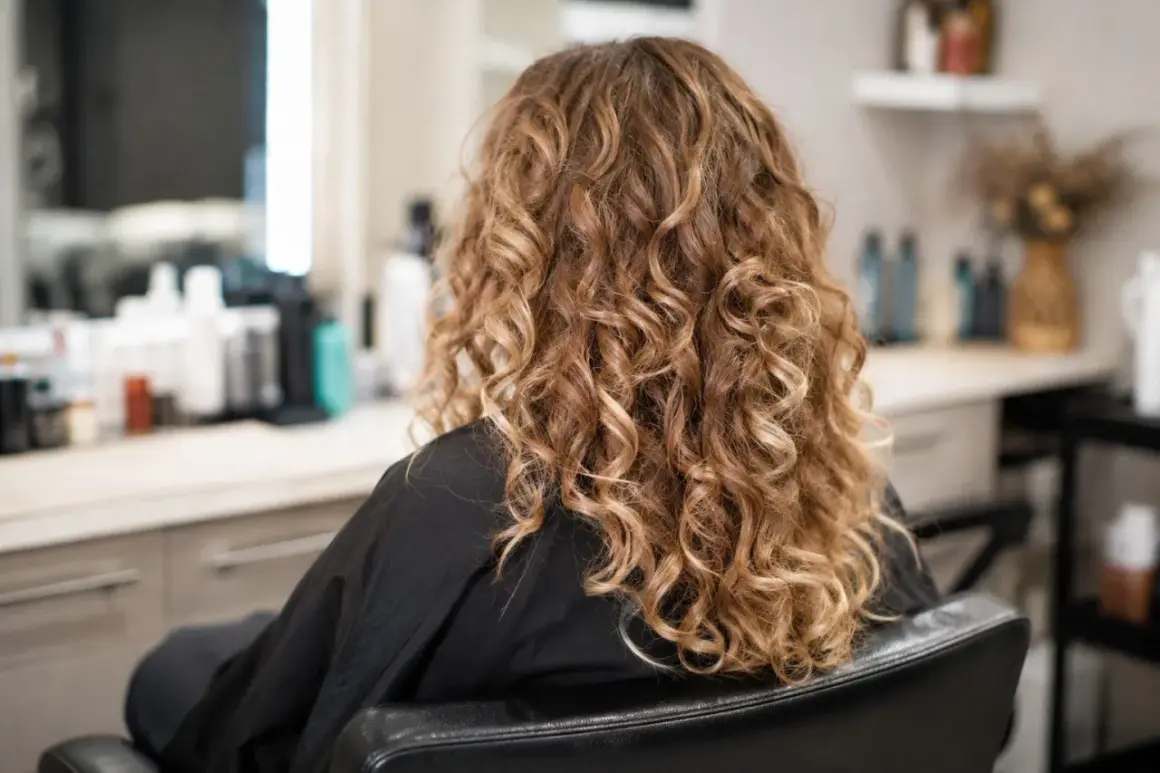 Kobieta w czarnym szlafroku, z pięknymi, złotymi loki trwała. W tle widać salon fryzjerski.