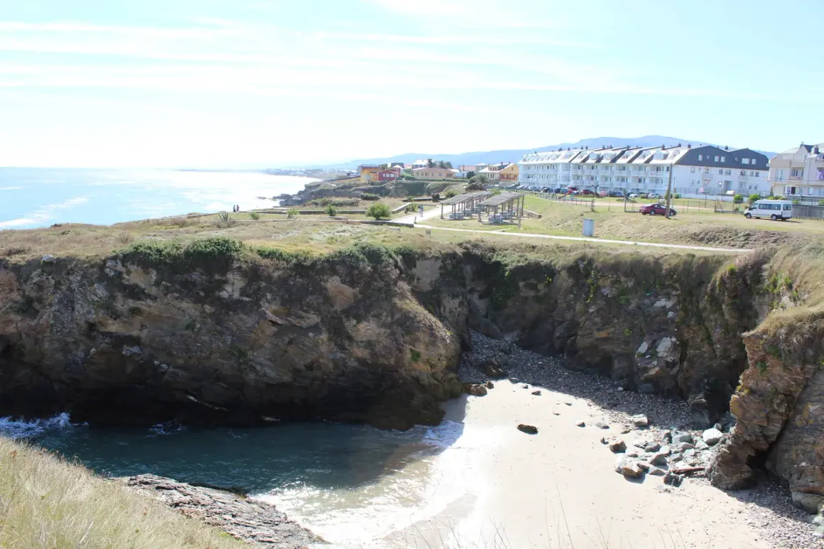 Acantilados rocosos y una pequeña cala de arena en la playa de San Bartolo Barreiros, con edificios residenciales al fondo.