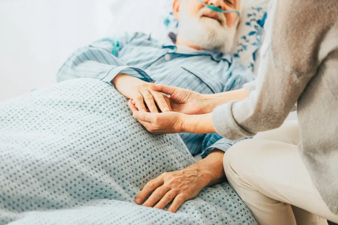 Ein älterer Mann mit Bart und Nasenbrille liegt im Bett. Eine Person hält seine Hand. Die Szene vermittelt die Endphase, wie man an Prostatakrebs stirbt.
