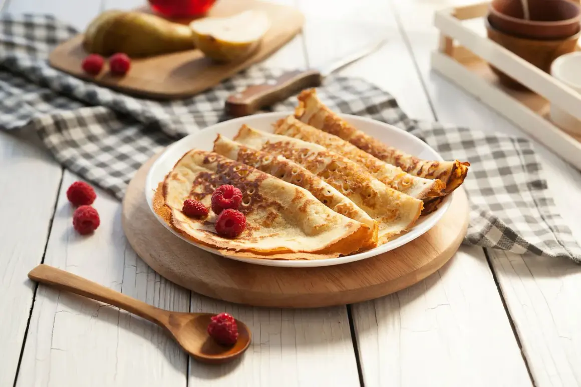 Frisch zubereitete Pfannkuchen, ein köstliches Pancakes Rezept, garniert mit Himbeeren auf einem Teller. Daneben liegen Birnen und ein Holzlöffel.