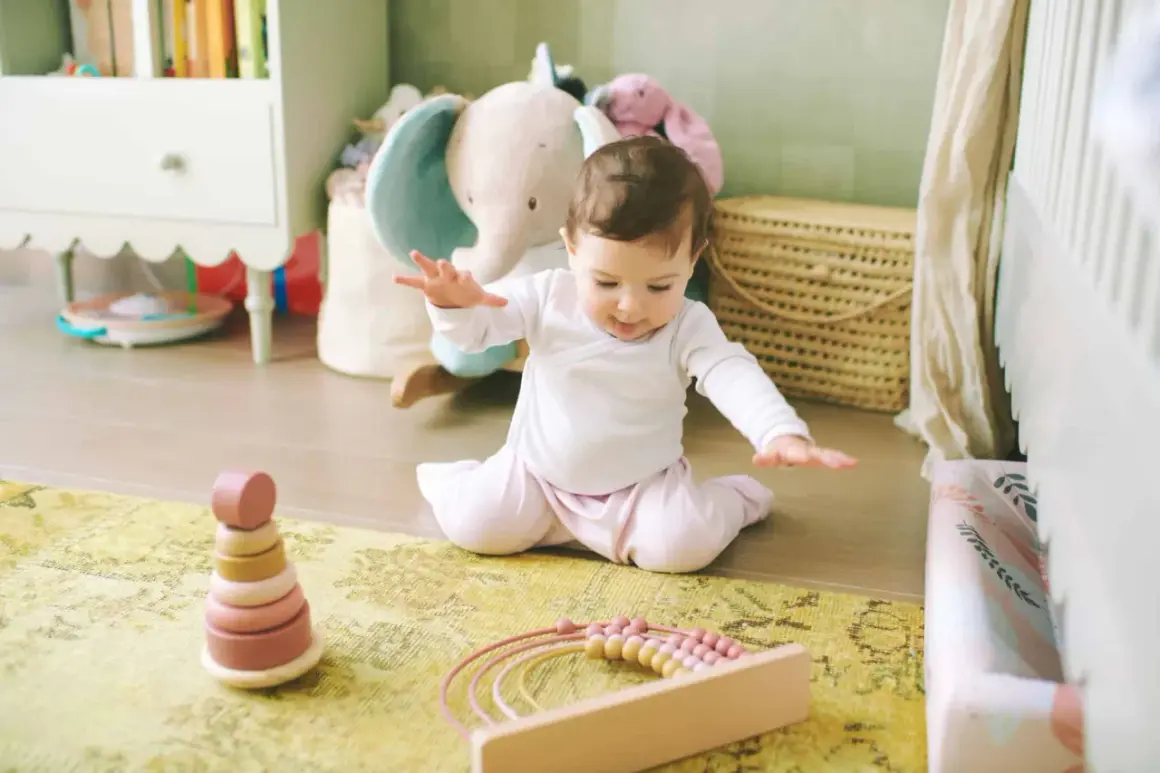Ein **baby 9 monate** sitzt auf einem Teppich und spielt mit Holzspielzeug. Im Hintergrund sind Stofftiere und ein Kinderbett zu sehen.