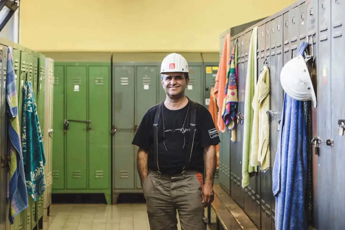 Ein Mann in Arbeitskleidung und Helm steht in einer Umkleidekabine. Handtücher hängen an den Spinden. Die Umziehzeit ist Teil der Arbeitszeit.