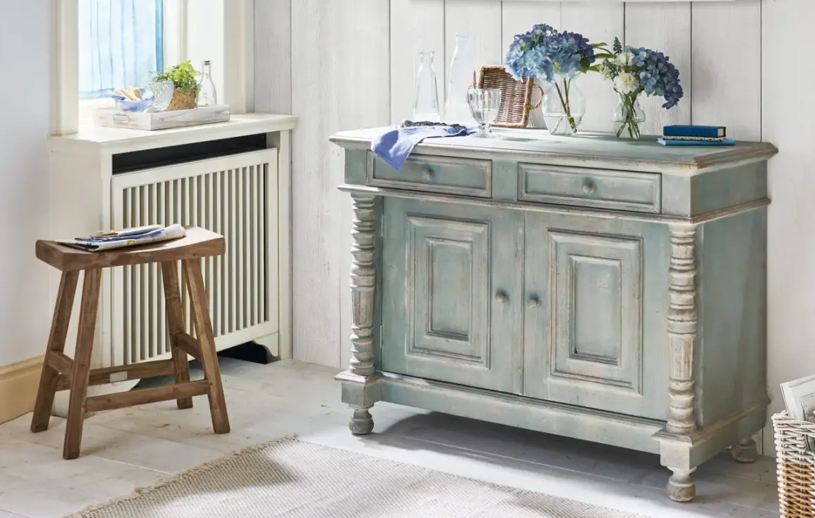 Ein blau-graues Sideboard mit abgenutzter Patina, das den "shabby chic"-Stil verkörpert. Daneben ein Holzhocker und Blumen.