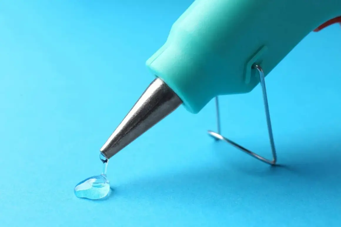 Un pistolet à colle turquoise dépose une goutte de colle transparente sur un fond bleu. Une petite tache de colle se forme.