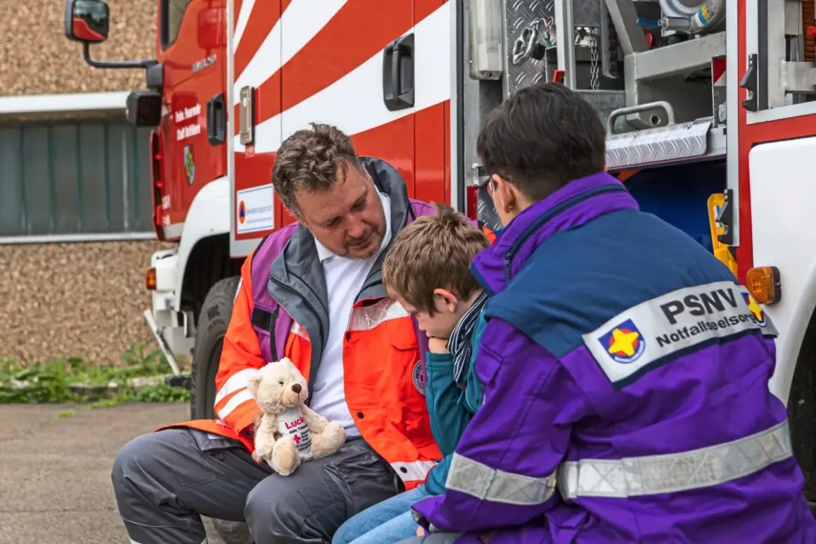 Ein Mann mit einem Teddybär spricht mit einem Kind neben einem Feuerwehrauto. Eine Person mit PSNV-Aufschrift auf der Jacke sitzt daneben.