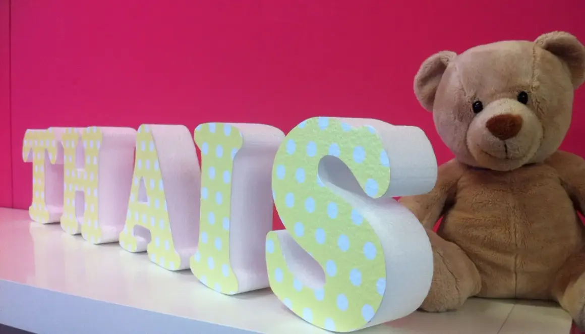 Lettre en relief "THAIS" décorée de pois sur fond rose vif, à côté d'un ours en peluche.