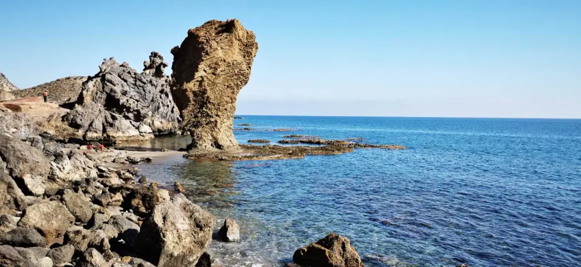 Rocas erosionadas y mar azul en la playa El Sombrerico. Una formación rocosa alta se alza como un guardián.