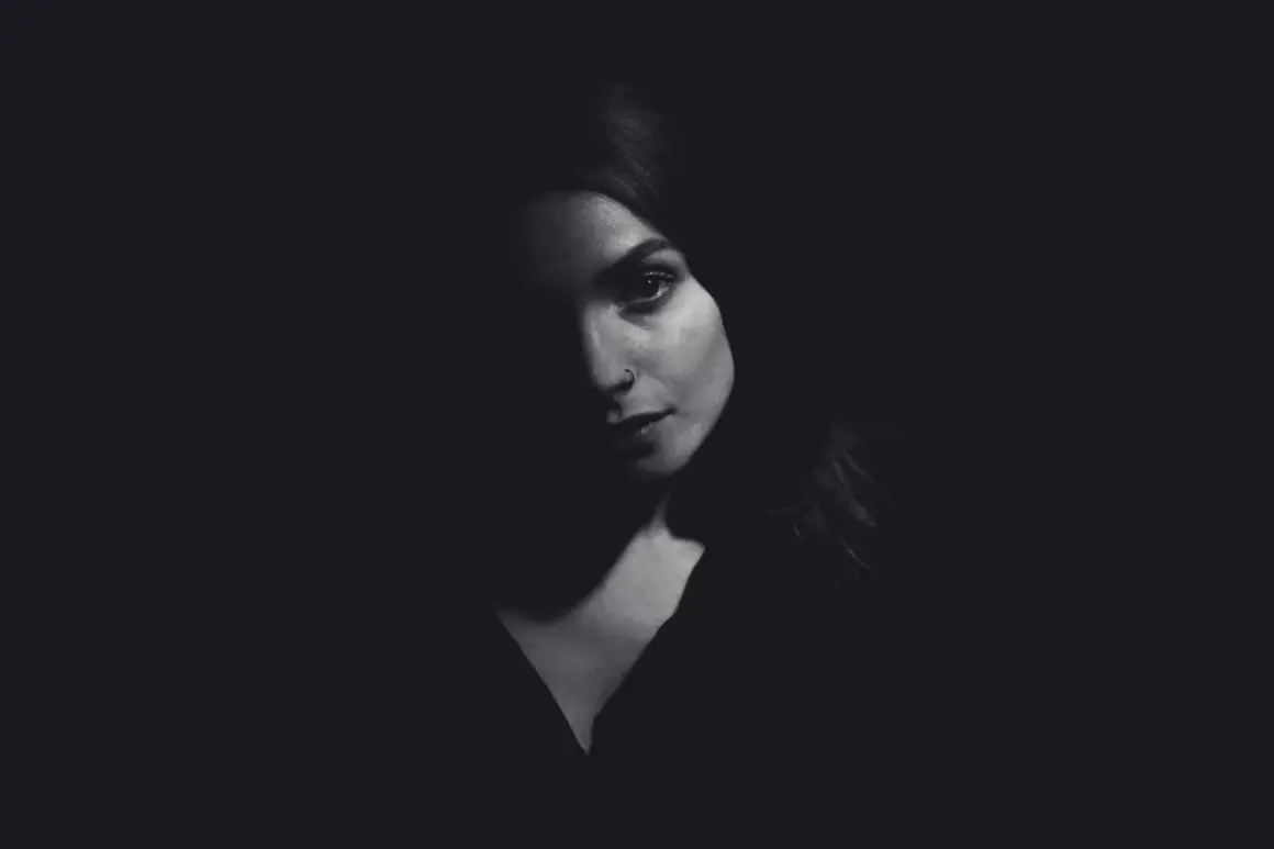 A woman's face emerges from darkness, her features dramatically sculpted by side lighting. A subtle nose ring catches the light.