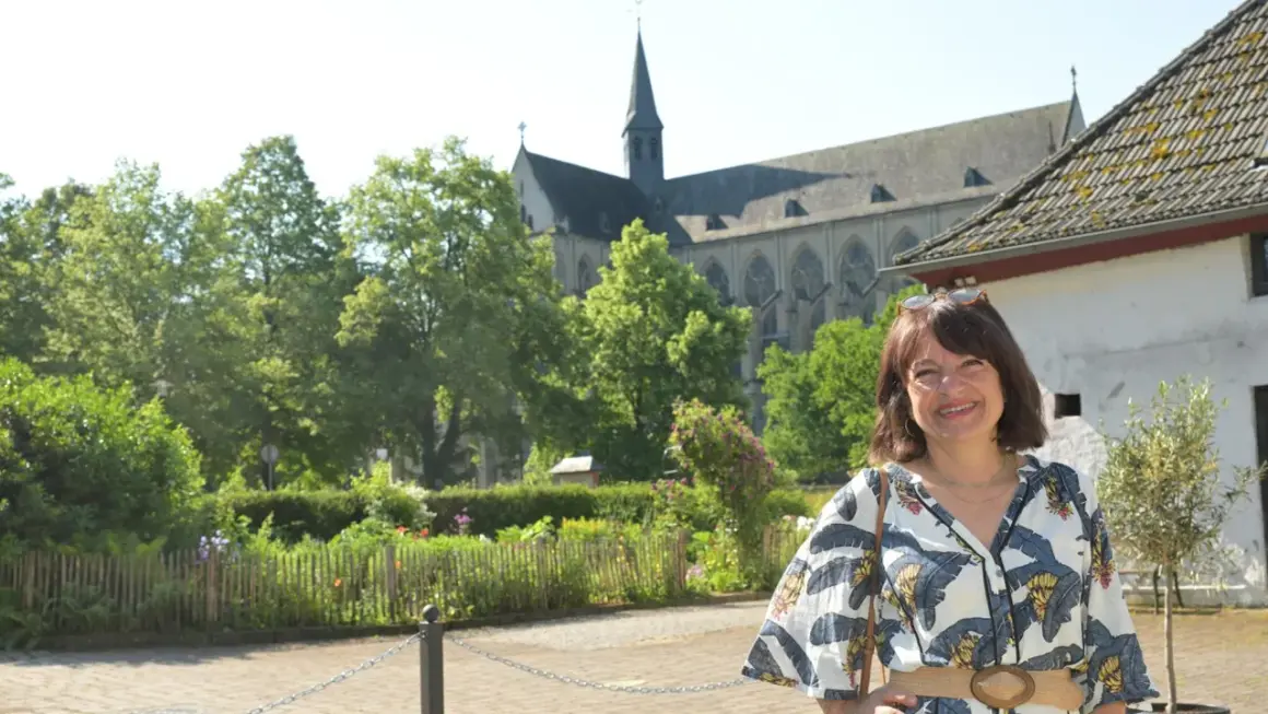 Frau lächelt vor einem Dom, umgeben von Bäumen und einem Garten. Ein Ort, der zum Verweilen und zur Entdeckung von Literatur am Dom einlädt.