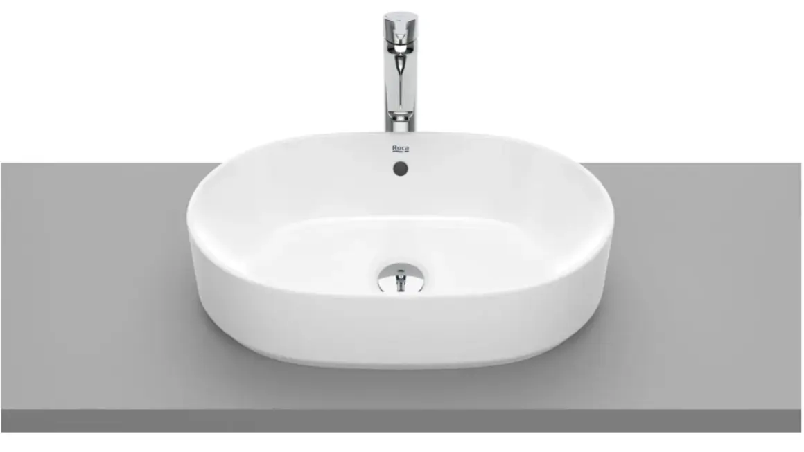 Elegancka, owalna umywalka nablatowa bez otworu na baterię, wykonana z białej ceramiki, z chromowanym korkiem.