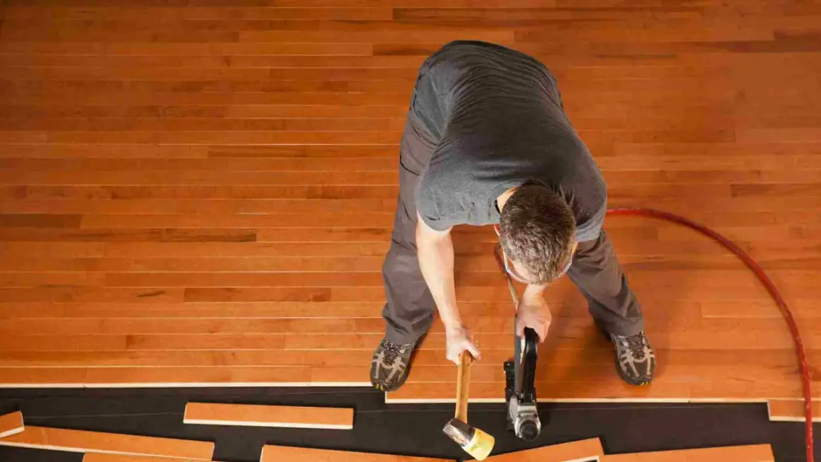 A man is installing waterproof flooring, using a nail gun and mallet to secure wooden planks.