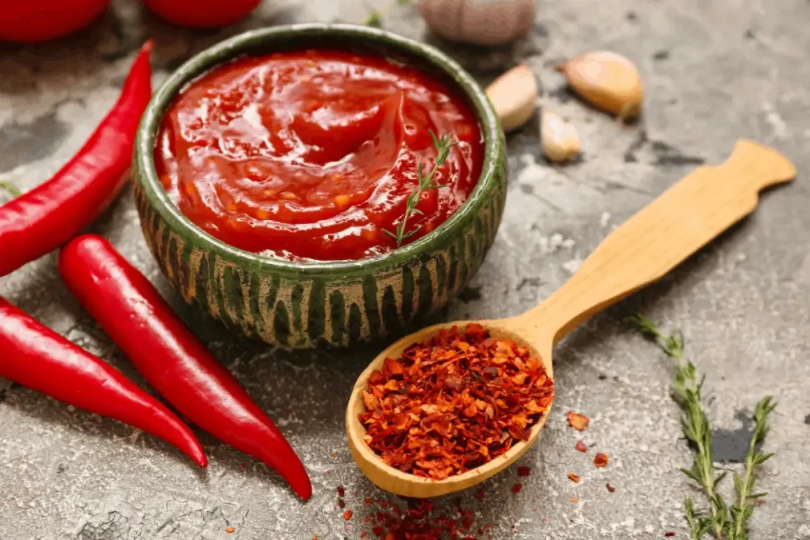 Miseczka pełna pikantnego sosu meksykańskiego, obok świeże papryczki chili i drewniana łyżka z płatkami chili.