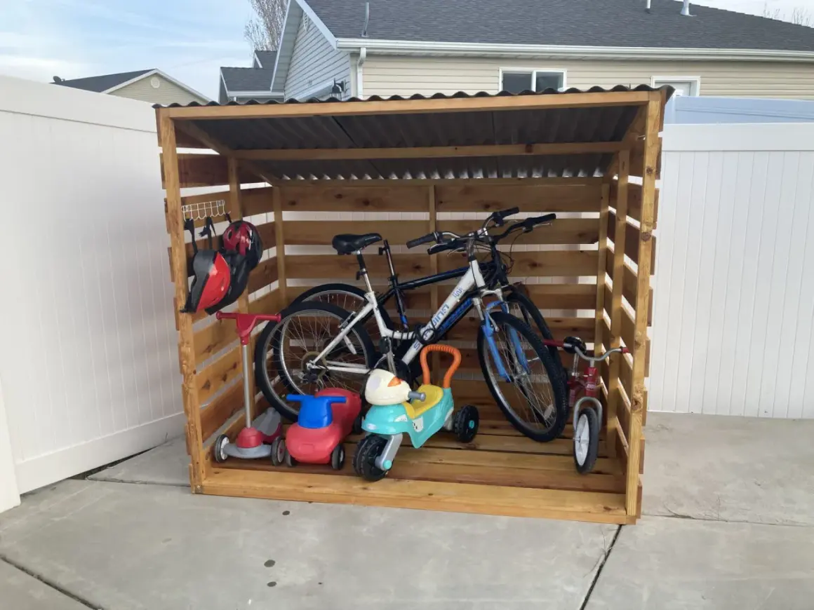 A DIY bike rack made of wood stores two adult bikes, a tricycle, and two ride-on toys. Helmets hang on a hook.