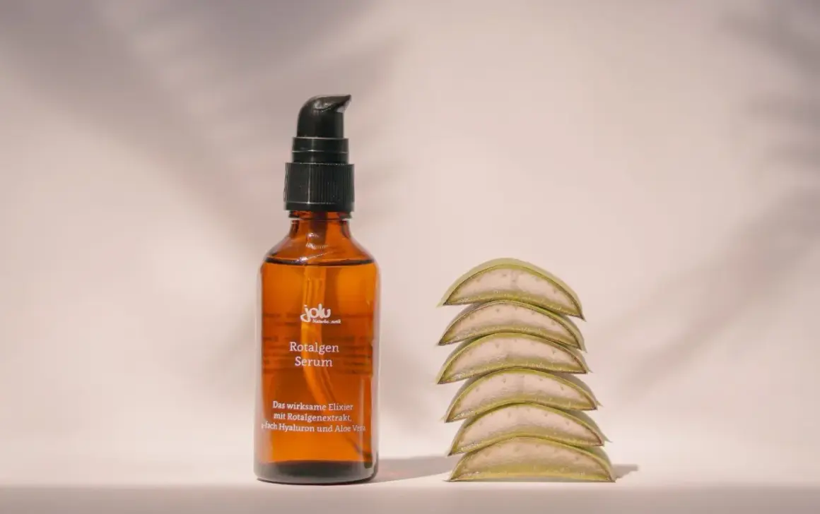 Braunes Fläschchen mit Rotalgen Serum, das Rotalgenextrakt als geliermittel enthält, neben aufgestapelten Aloe-Vera-Scheiben.