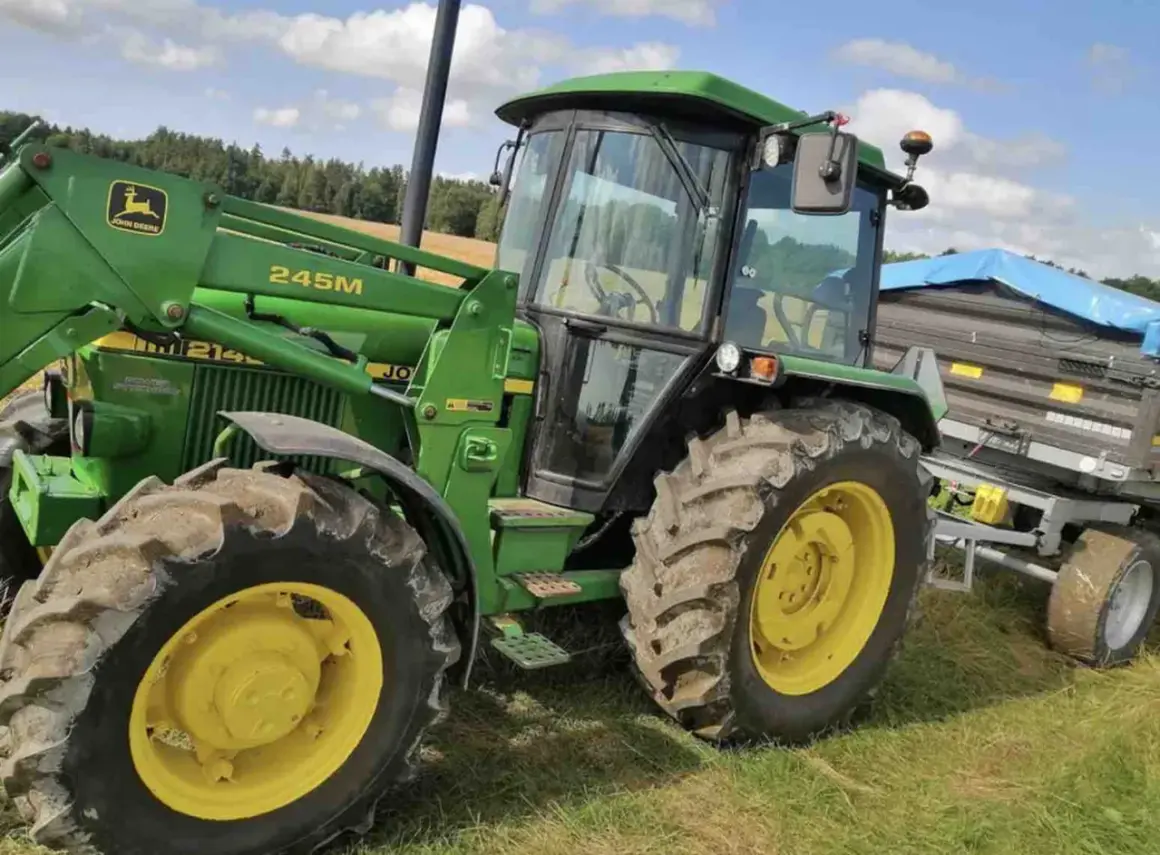 Zielony traktor John Deere 2140 z ładowaczem czołowym i przyczepą na tle łąki i lasu.