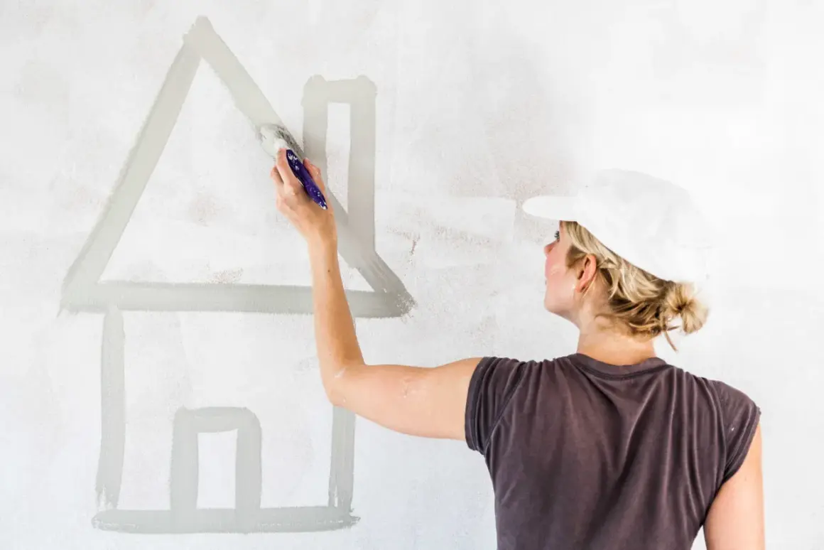 Donna con cappellino bianco dipinge una casa stilizzata su muro. Calcolare quanti litri di pittura per 50 mq.