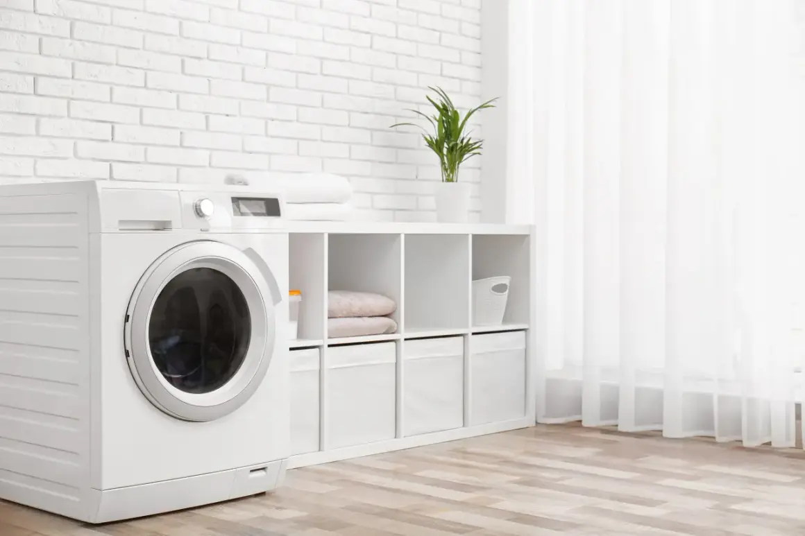 Weiße Waschmaschine mit beladener Trommel steht neben einem Regal mit Handtüchern und Aufbewahrungsboxen. Die Waschmaschine wandert nicht, sie steht fest.
