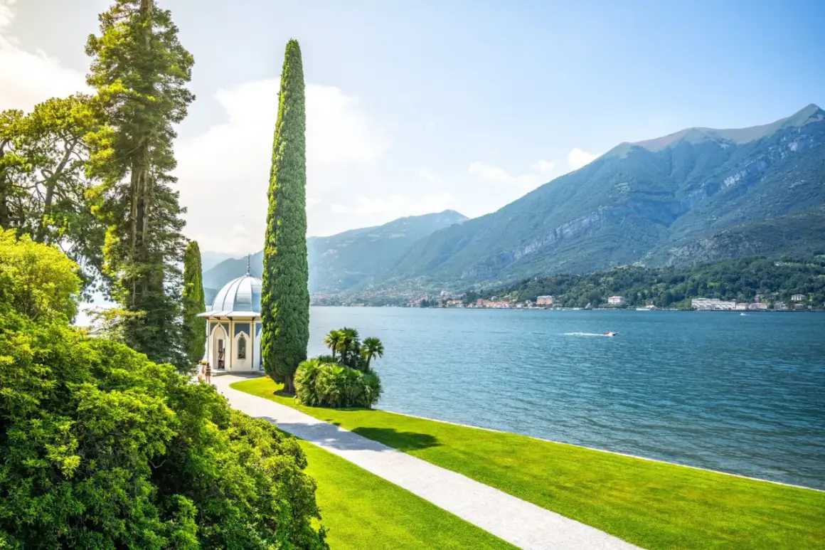 Giardino lussureggiante con gazebo bianco e cipressi slanciati, affacciato sul blu intenso del lago di Como, con montagne verdi sullo sfondo.