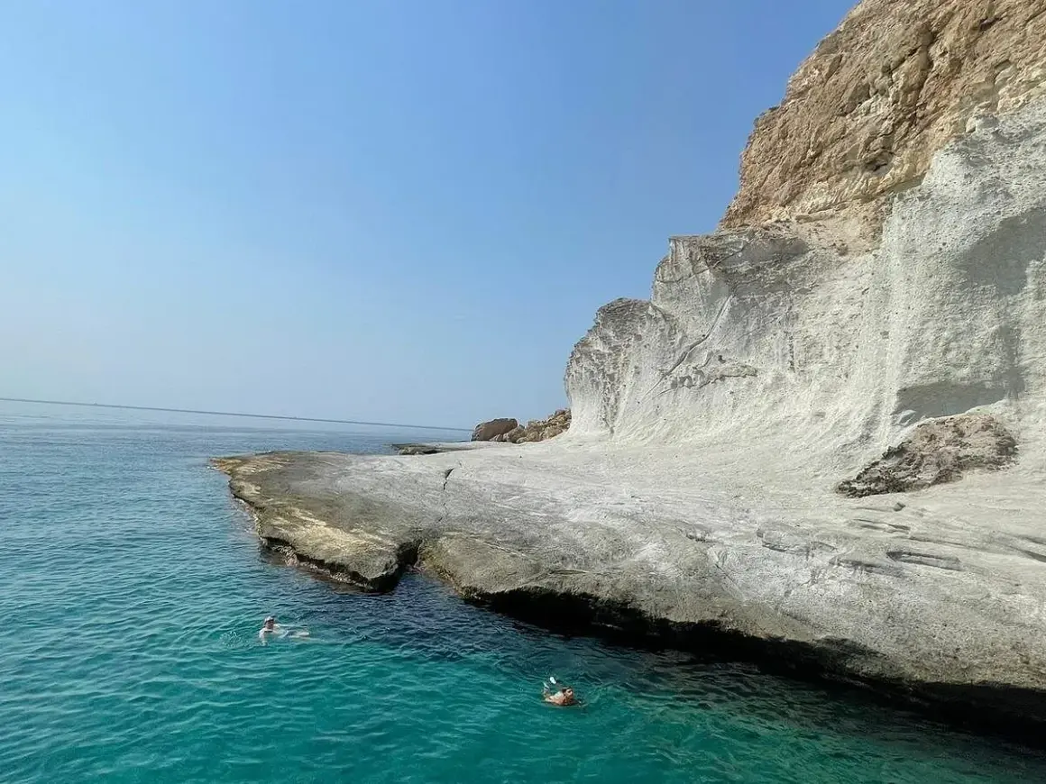 Dos personas nadan en las **calas agua amarga**, con acantilados blancos y mar turquesa bajo un cielo azul.