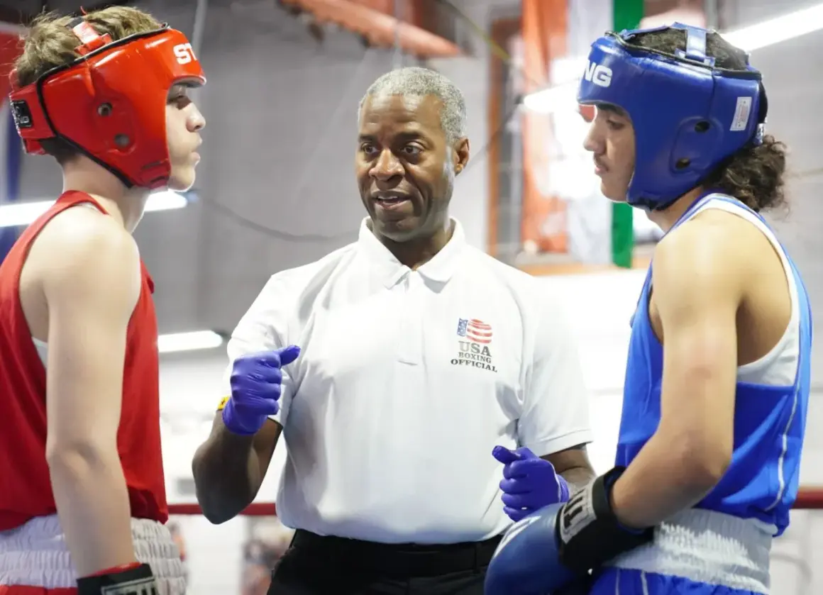 Referee instructs two young boxers in red and blue headgear during an amateur boxing match.