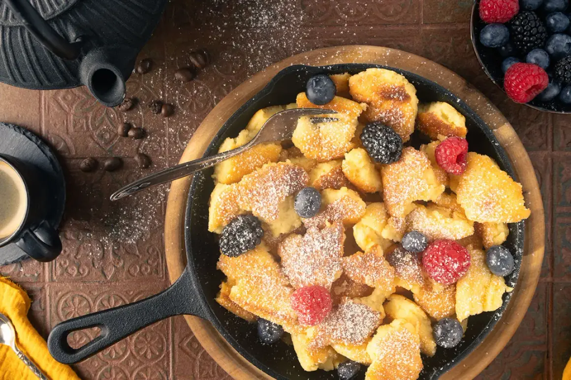 Ein herzhafter gerupfter Pfannkuchen, bestreut mit Puderzucker und garniert mit frischen Beeren, serviert in einer Gusseisenpfanne.