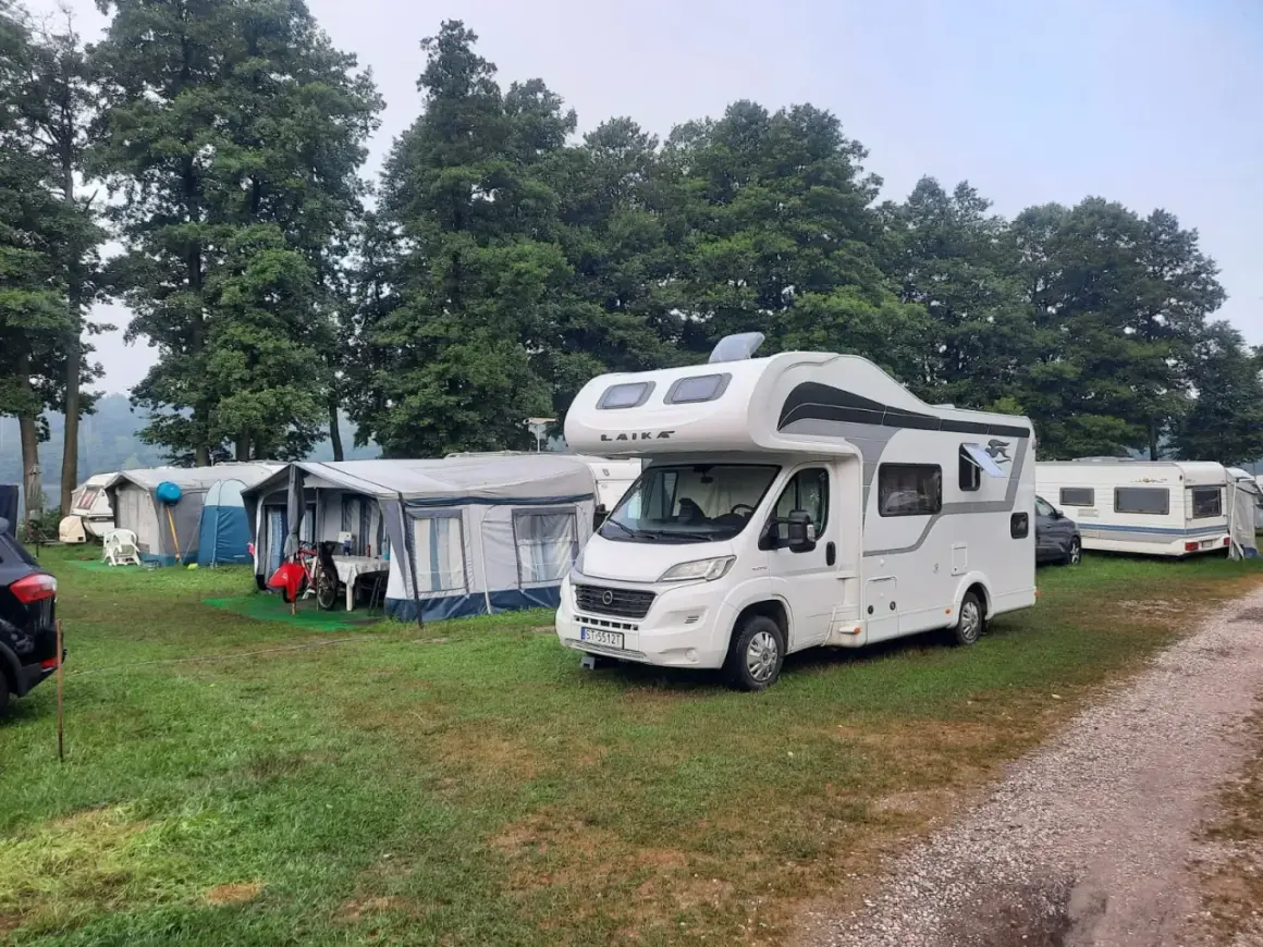 Kampery i przyczepy kempingowe na trawiastym polu, w tle drzewa. Widać namioty i meble ogrodowe. To idealne miejsce na wypoczynek w camp park sonata.