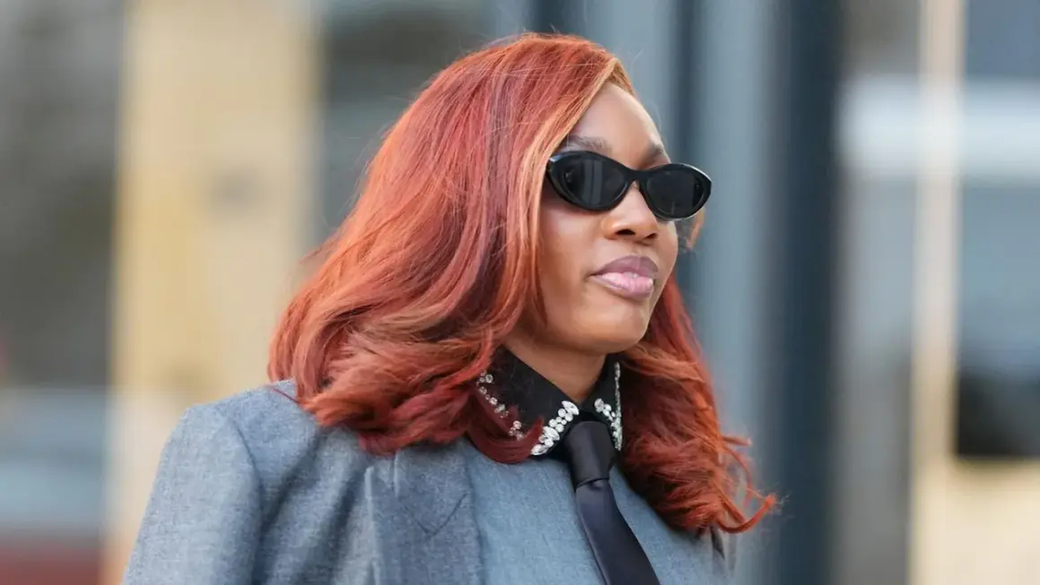 Frau mit feuriger, **cooler Haarfarbe** und stylischer Sonnenbrille trägt einen grauen Blazer mit Krawatte und glitzerndem Kragen.