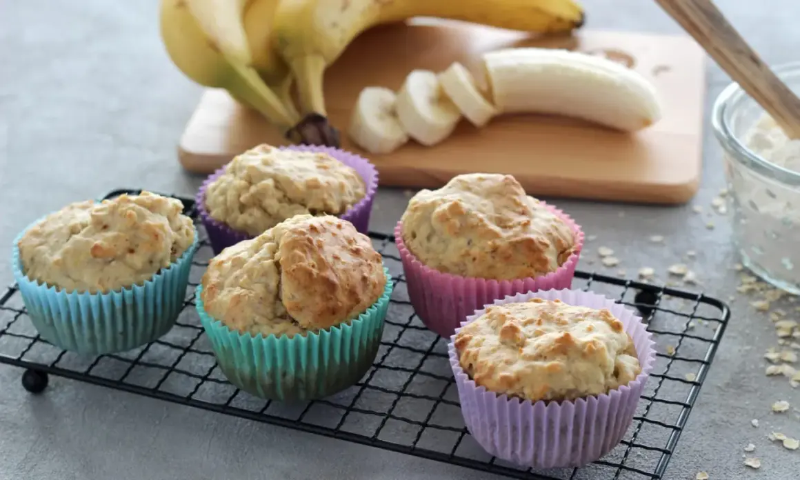 Fünf leckere Muffins ohne Zucker in bunten Förmchen, daneben Bananen und eine Schüssel Mehl.