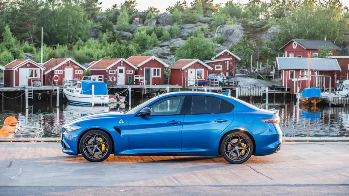 Blå sportbil vid en svensk hamn med röda sjöbodar. Undrar hur långt har bilen gått på denna vackra resa?