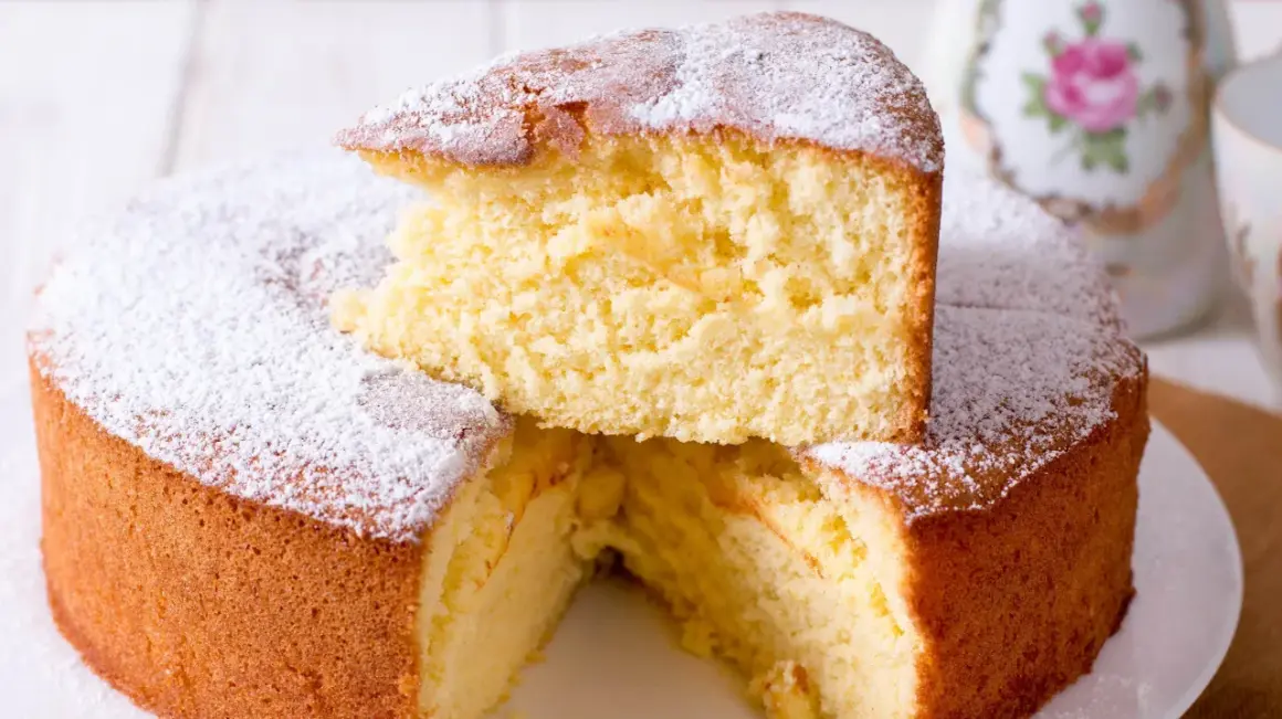 En fluffig vegansk sockerkaka, pudrad med florsocker. En bit är upplagd ovanpå resten av kakan.