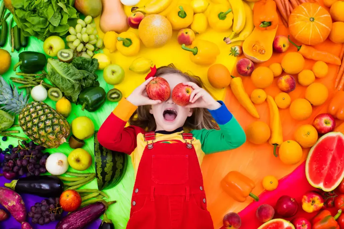 Enfant joyeux avec des pommes devant les yeux, entouré de fruits et légumes colorés. Une belle scène pour une activité manuelle semaine du goût.
