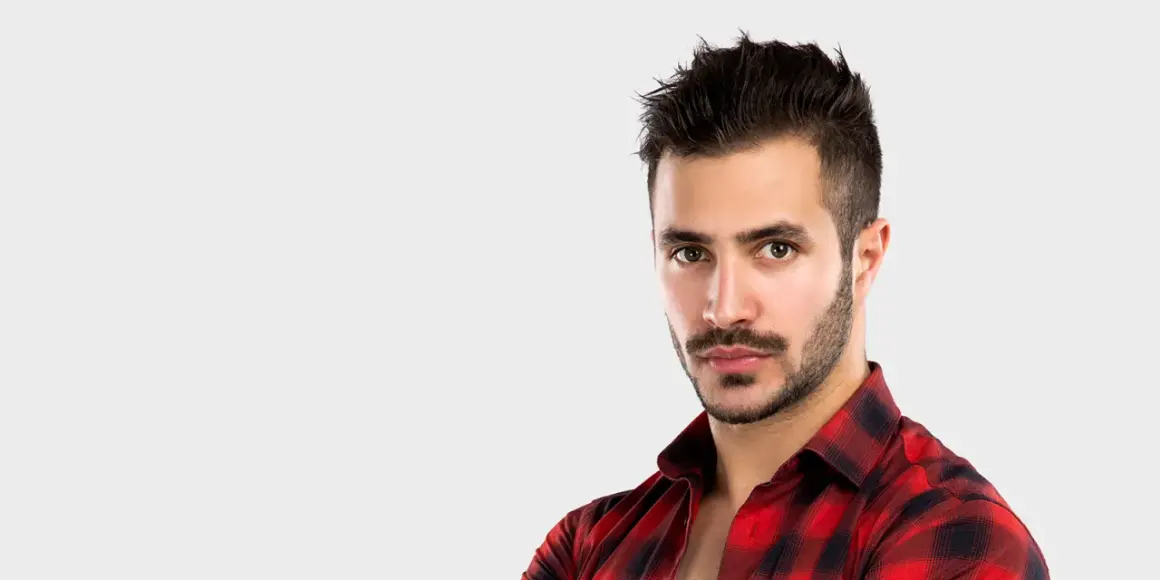 Un homme au regard intense, avec une barbe mouche bien taillée, porte une chemise à carreaux rouges et noirs.