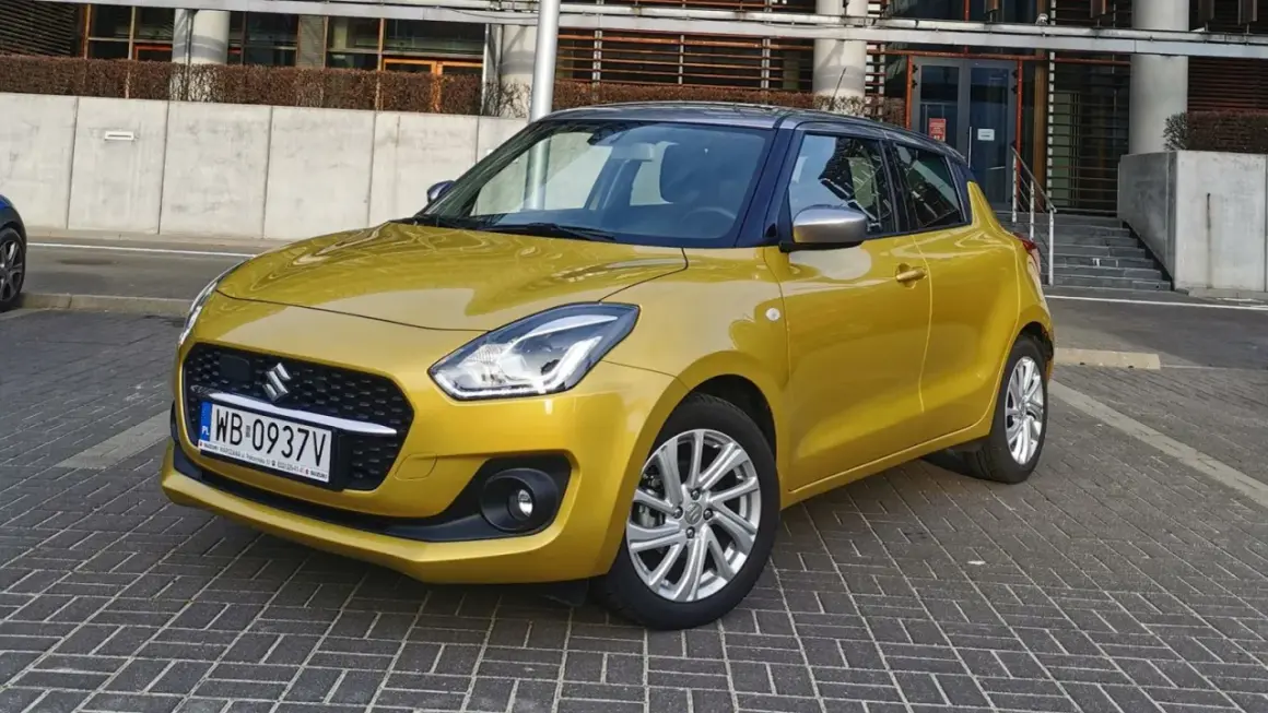 Złoty Suzuki Swift na tle nowoczesnego budynku. Wiele pozytywnych suzuki swift opinie potwierdza jego miejski charakter.