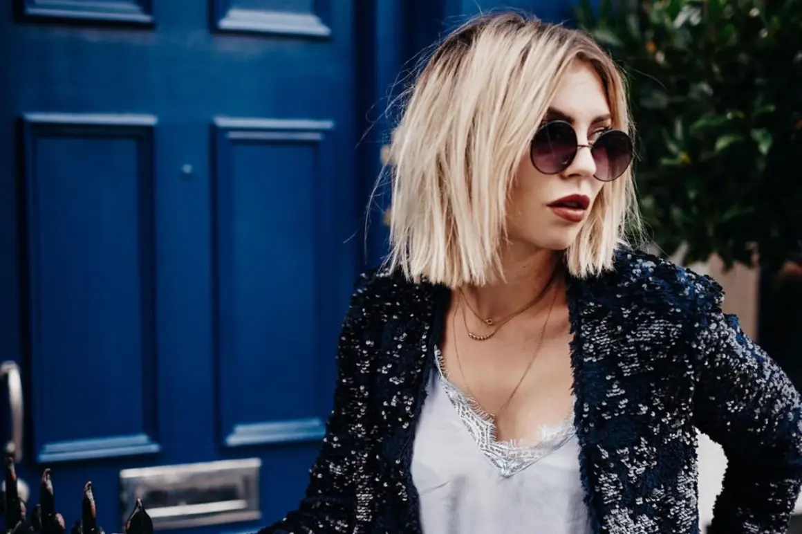 Frau mit lässiger hipster frisur, Sonnenbrille und Paillettenjacke vor blauer Tür.