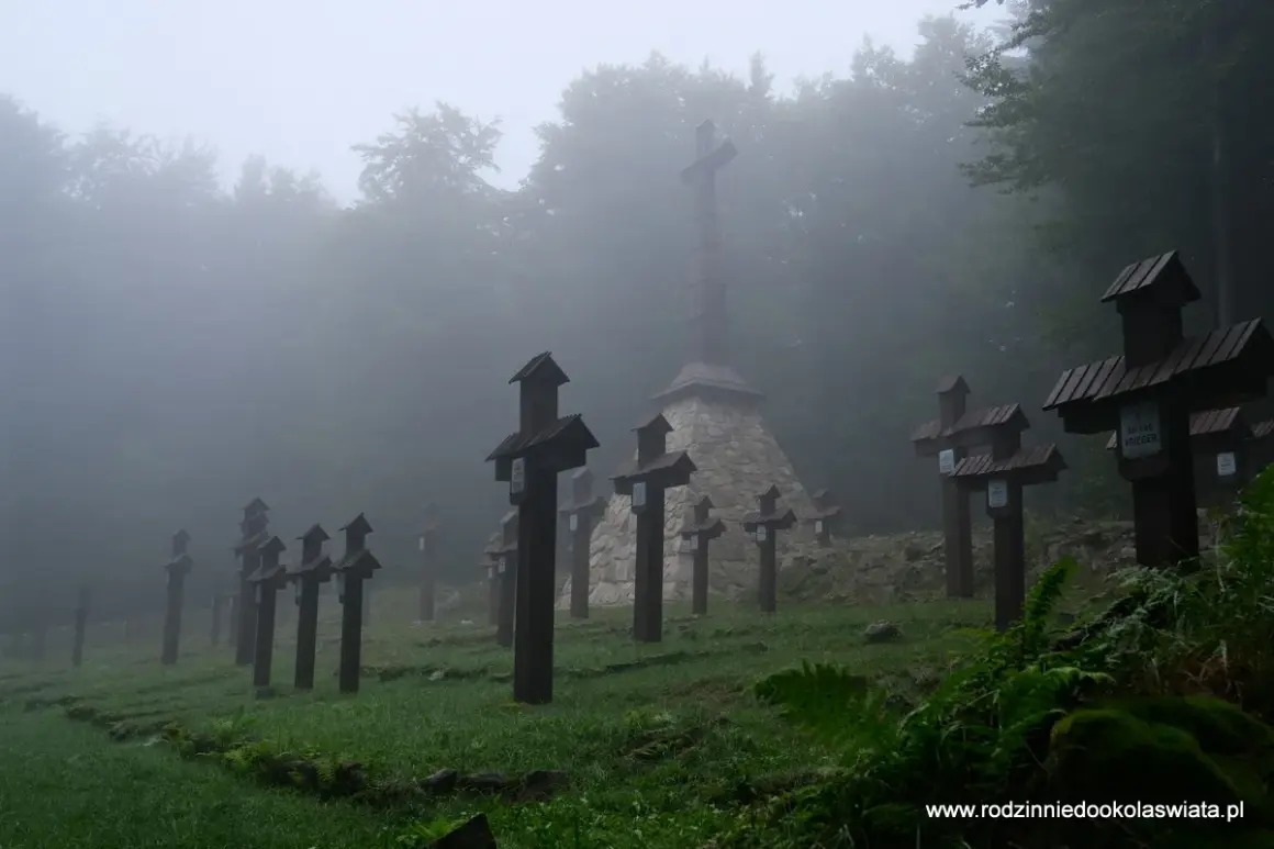 Mglisty poranek na cmentarzu wojennym na Magurze Malastowskiej. Drewniane krzyże stoją w szeregach, a w tle wznosi się monumentalny pomnik.