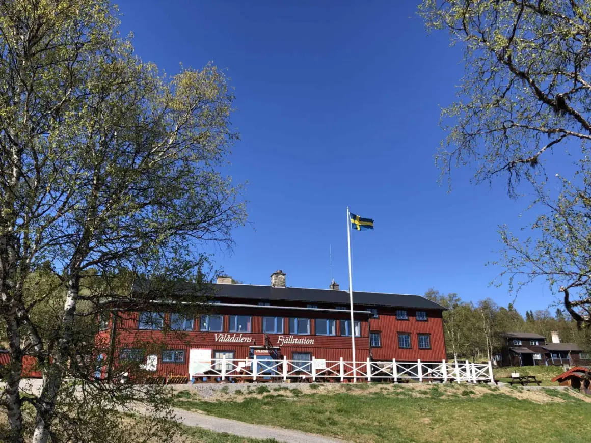 STF Vålådalen fjällstation i solsken med svensk flagga. Träd med gröna blad ramar in den röda byggnaden.