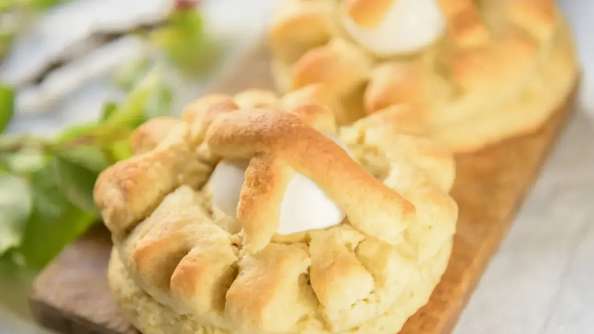 Dolci pasquali pugliesi con uova intere, decorati con impasto intrecciato, su un tagliere di legno.