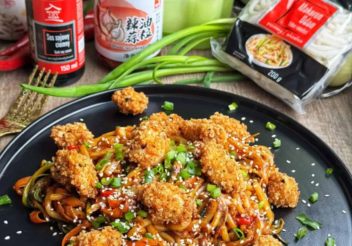 Pyszne udon z kurczakiem w panierce, posypane sezamem i szczypiorkiem. Obok sos sojowy i opakowanie makaronu udon.