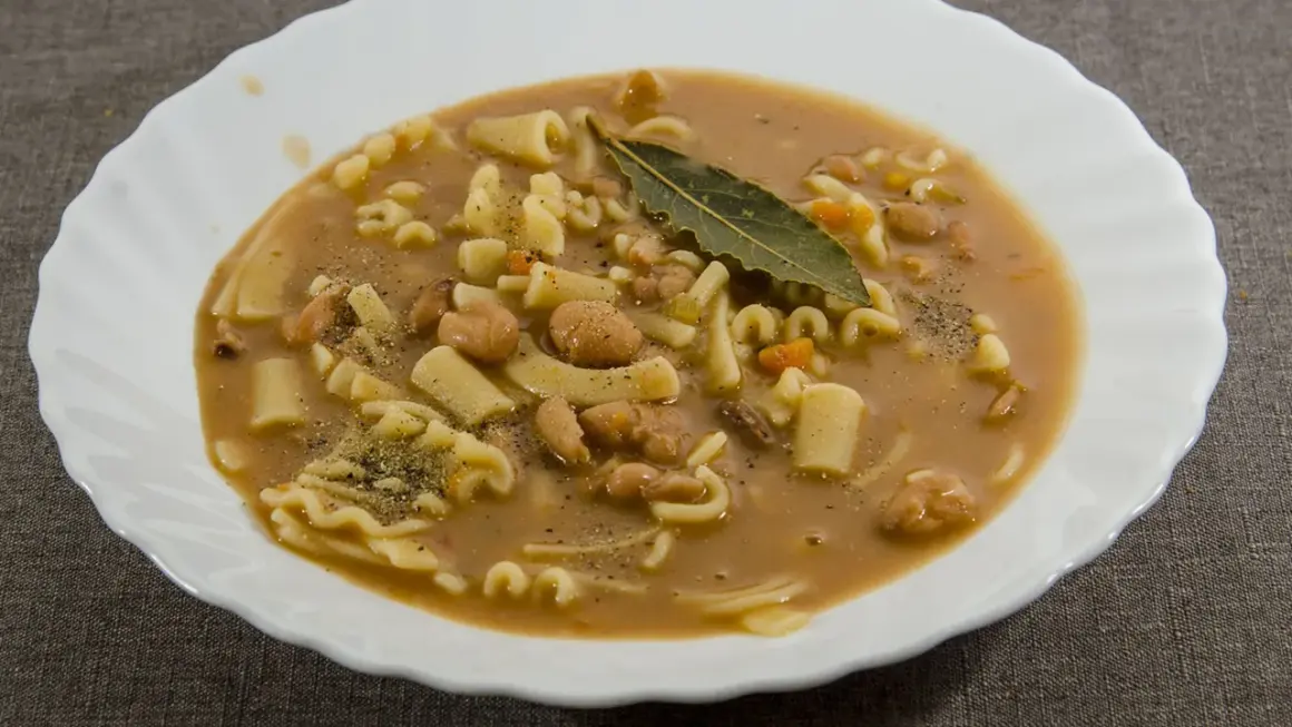 Un piatto fumante di pasta e fagioli, con pasta corta, fagioli borlotti, carote e una foglia di alloro.