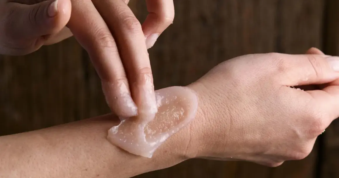 Mani che applicano scrub corpo sulla pelle, mostrando come si usa lo scrub corpo per una pelle liscia e luminosa.
