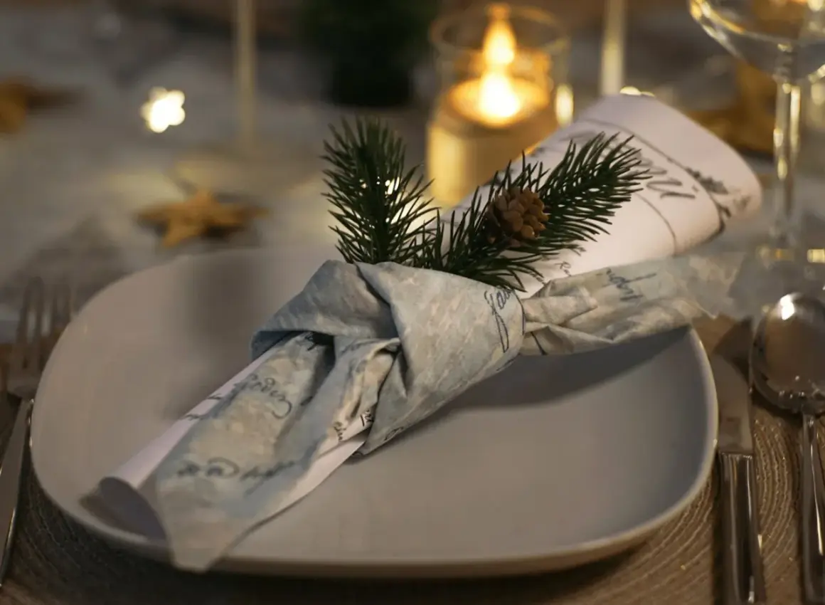 Une serviette en tissu, décorée d'une branche de sapin et d'une pomme de pin, est pliée en forme de smoking sur une assiette.