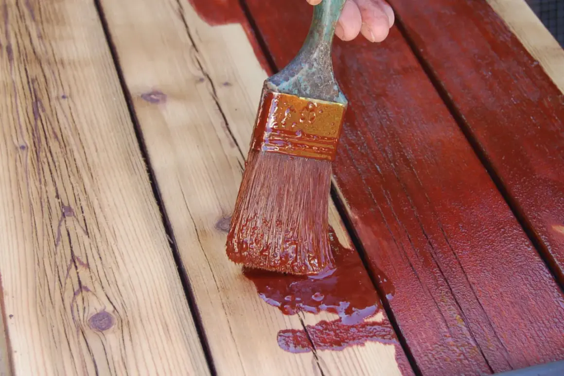 Application d'une teinture rouge sur des planches de bois. Un pinceau chargé de produit montre comment peindre du bois pour le protéger.