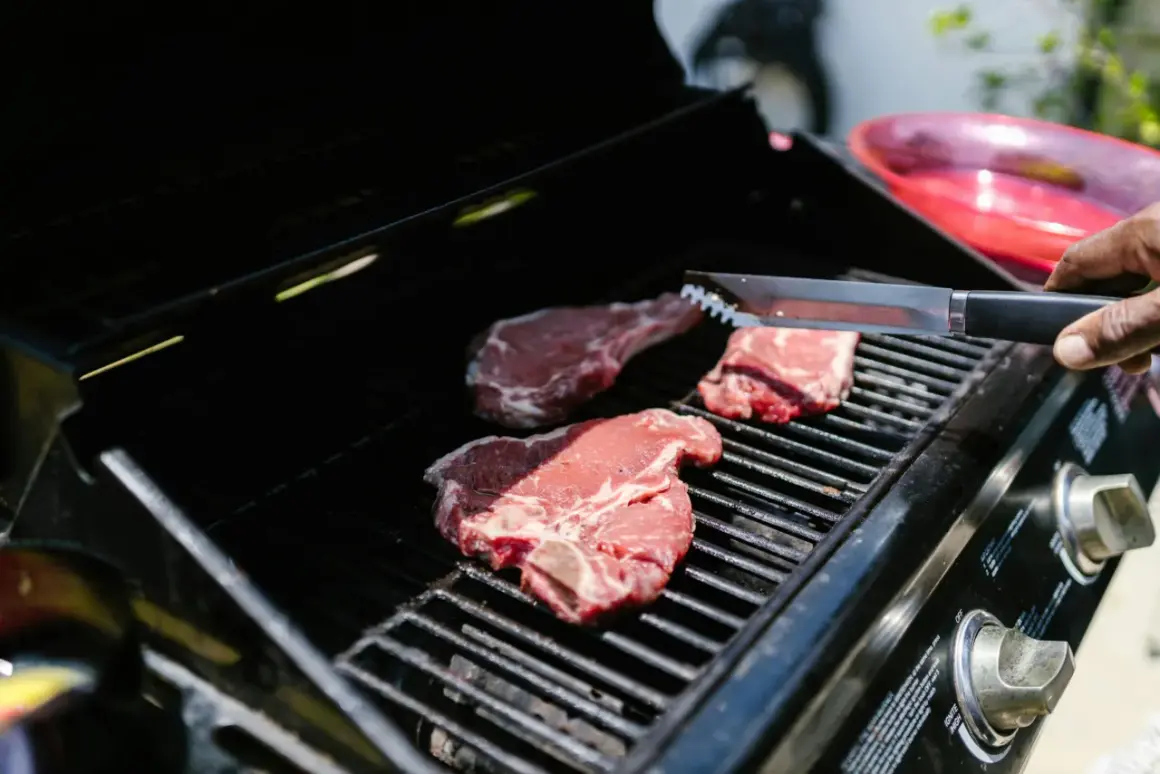 Trzy soczyste steki na ruszcie grilla, gotowe do grillowania. Dłoń z szczypcami zbliża się do mięsa.