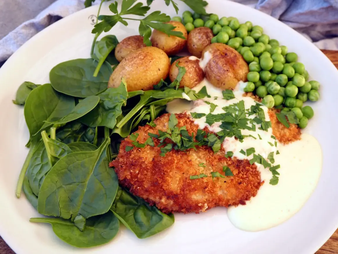 En panerad schnitzel med krämig sås, serverad med färsk spenat, små potatisar och gröna ärtor. En klassisk rätt där såsen till schnitzel lyfter hela smakupplevelsen.